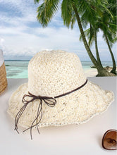Load image into Gallery viewer, A woven white hat with brown string bow is shown on a beach backdrop.
