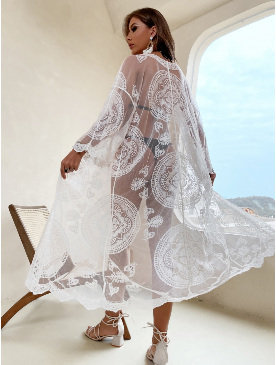 A woman wearing a white kimono with a lace pattern is standing in a white room. You can see through the kimono slightly and see a black bikini.