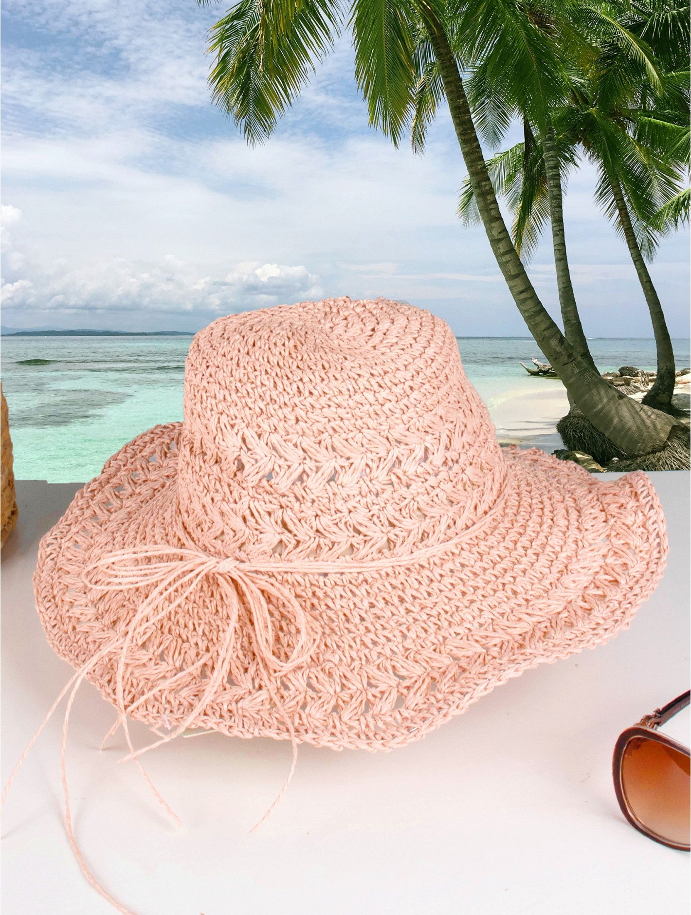 A woven pink hat with pink string bow is shown on a beach backdrop.