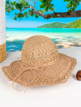 Load image into Gallery viewer, A woven light brown hat with light brown string bow is shown on a beach backdrop.
